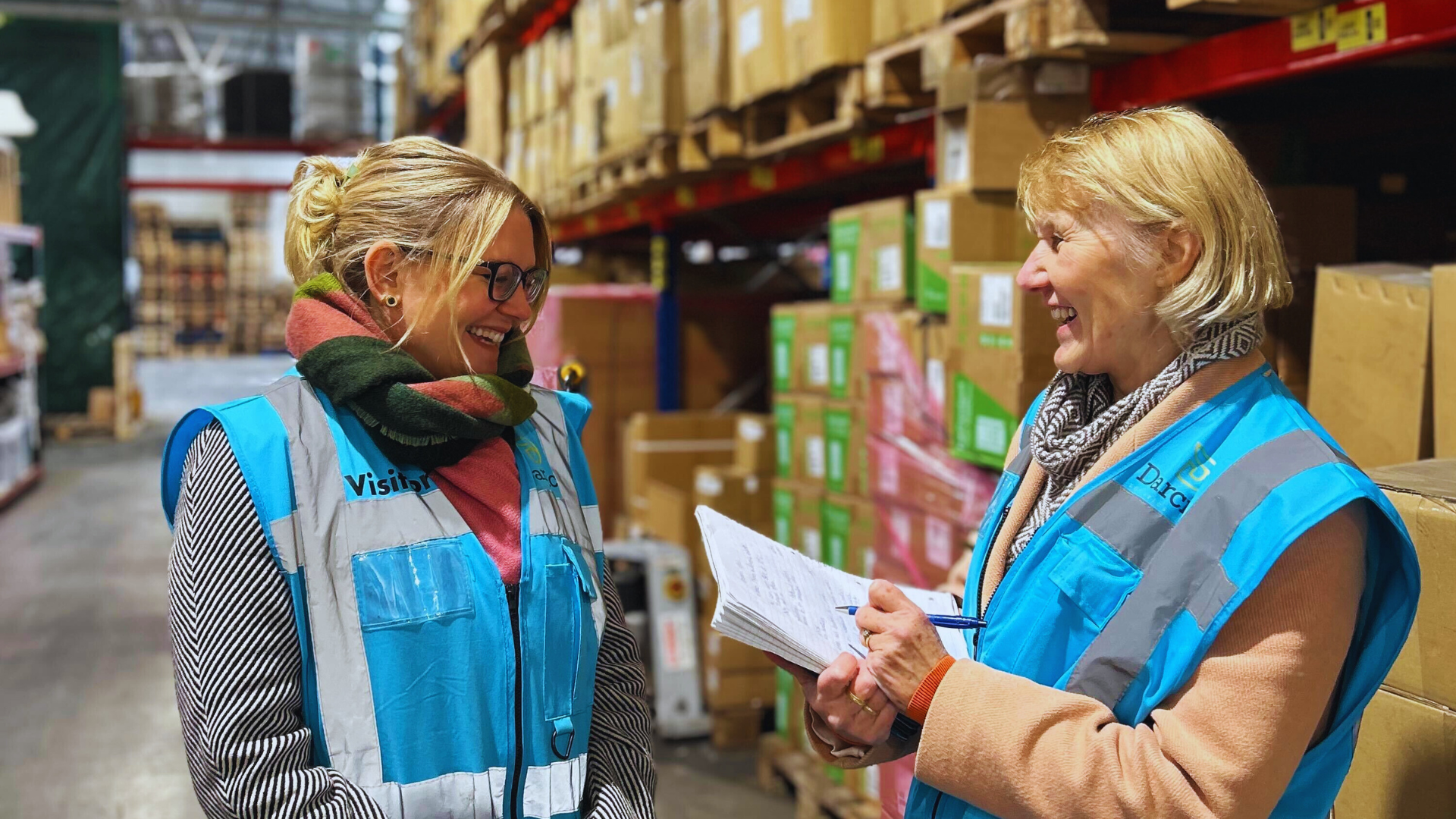 Two women stand in a warehouse full of brown cardboard boxes, wearing blue high-vis jackets and smiling widely at each other.