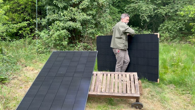 A man in a leafy green clearing carries a solar panel towards an empty space in a structure on the ground that is already holding up a second solar panel at an angle.