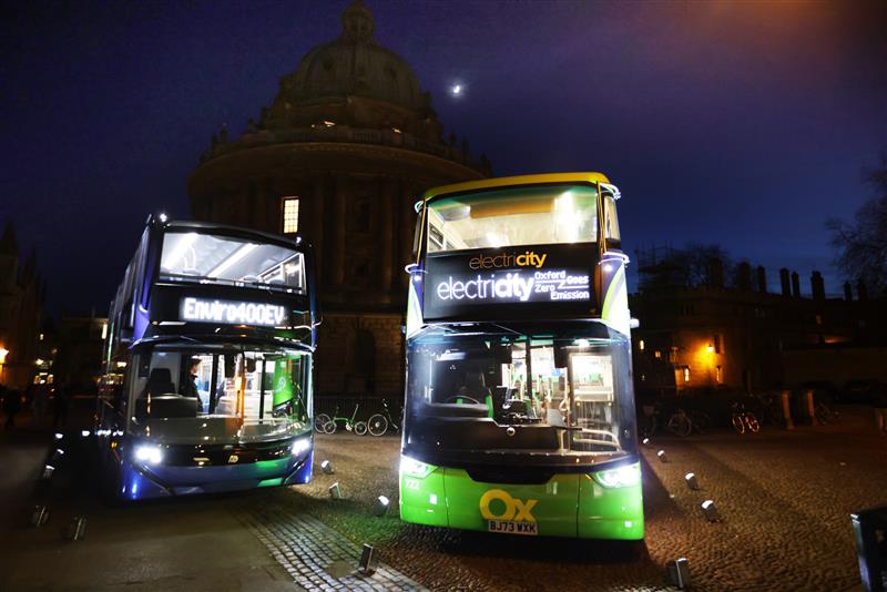 Two double decked buses sit in the road facing towards the camera. It is night time and their lights are shining strongly. Electric text on the buses displays read 'electri-city'.