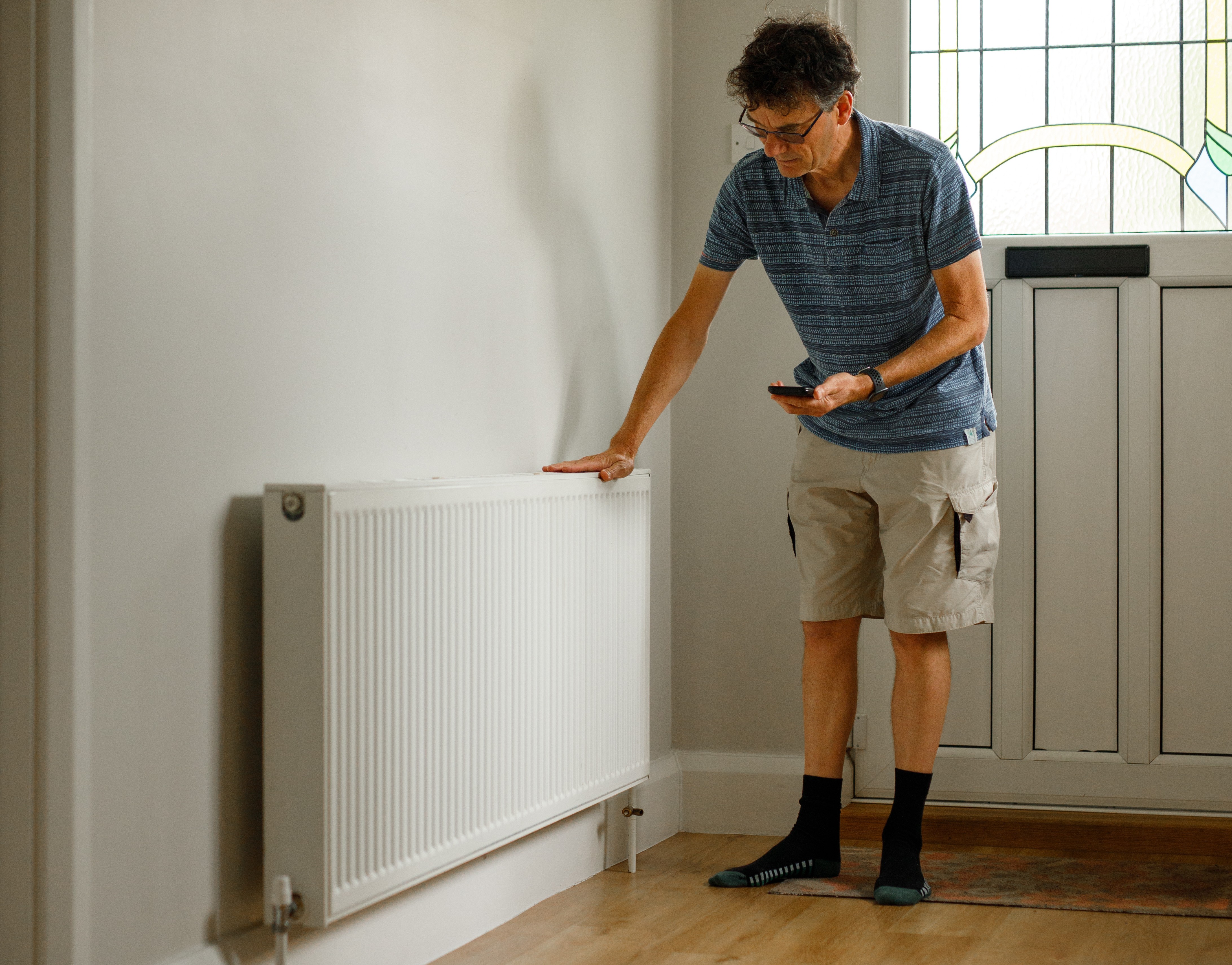 A man stands in his hallway with one hand pressed against a radiator and one hand holding a smartphone.
