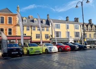 Six different-coloured EV cars line up in a town square on a sunny day, vertical banners flying next to each one.