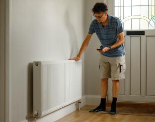 A man stands in his hallway with one hand pressed against a radiator and one hand holding a smartphone.