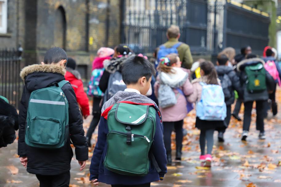 A group of primary school age children walk along a pavement with their teacher.