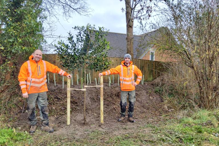 Two smiling men in orange high vis standing next to a tree the planted in a park next to a house fence