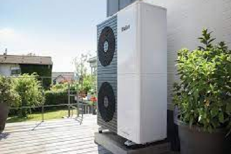 A white and grey air source heat pump on a deck next to a wall near a garden.