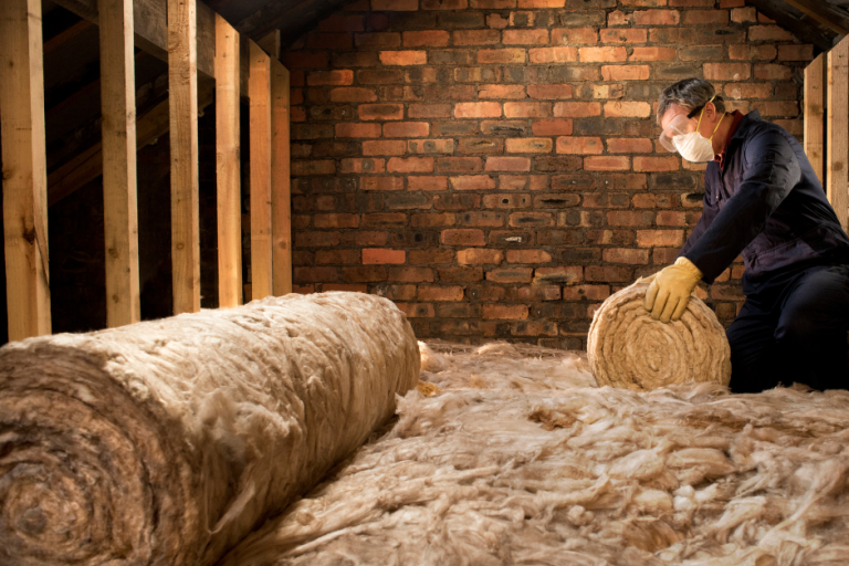 A man wearing a mask an blue overalls rolling out insulation in a warmly lit attic