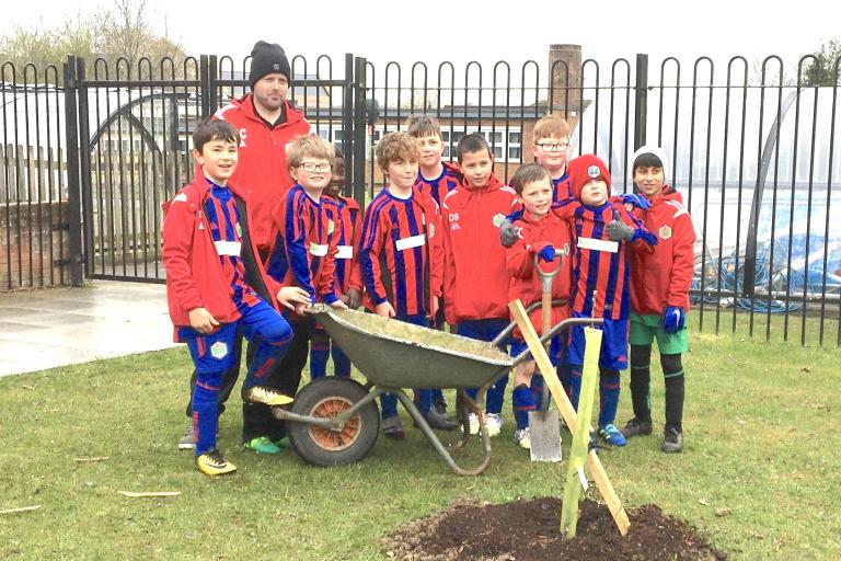 A football team of young boys smiling in front of a newly-planted tree