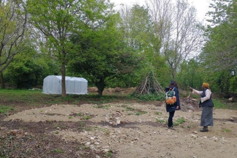 An empty plot of land surrounded by trees with two women standing in it. A greenhouse and stick fort can be seen in the background.