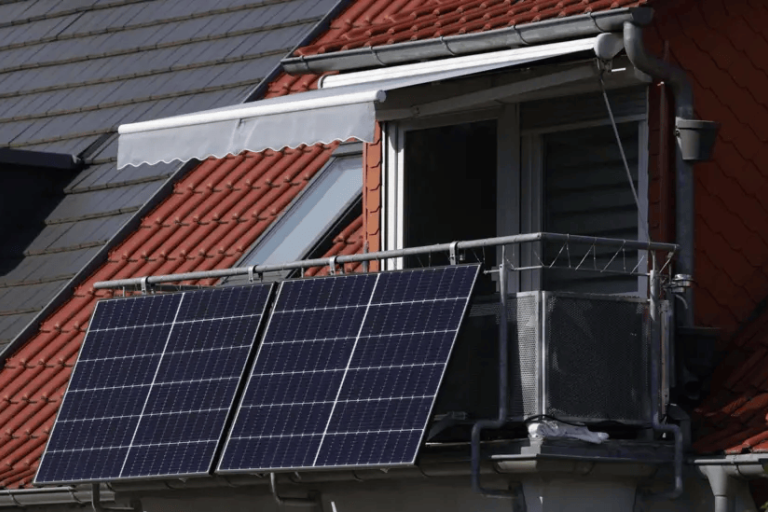 A red tiled, slanted rooftop containing a balcony with four slanted solar panels attached to its exterior.