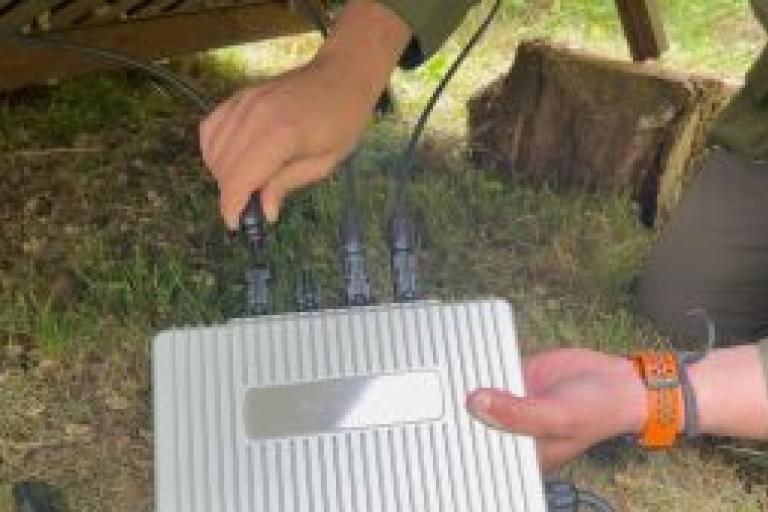 Two hands hold up an inverter against the grass, plugging a wire into the silver box.