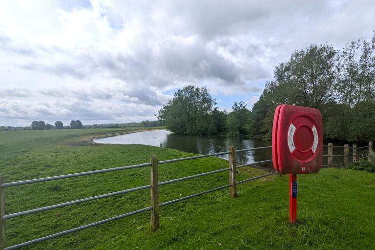The river Thames and it's grassy embankments, with a bright orange life ring post standing next to some metal fencing by the water.