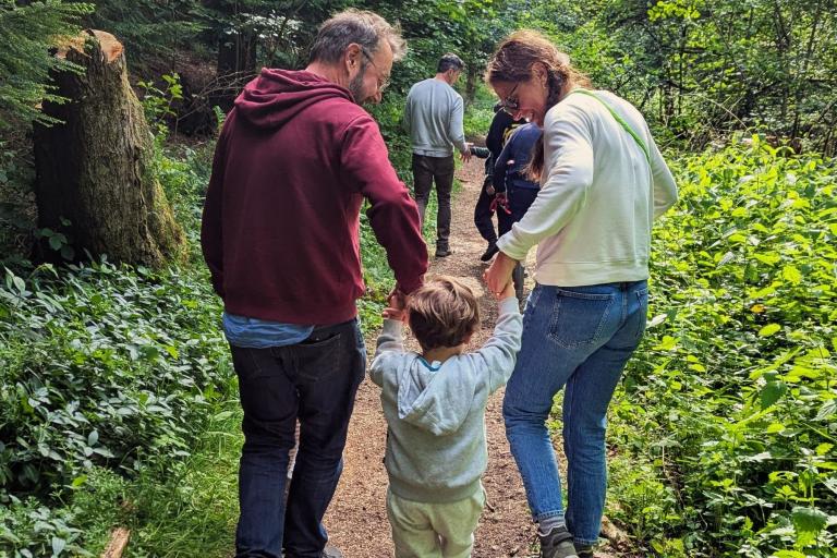 A group of people, including a mother, father, and child holding hands, walk along a woodland path.