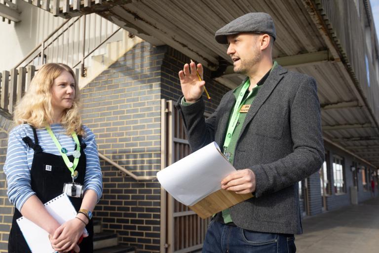 A woman and a man stand outside a school with clipboards chatting.