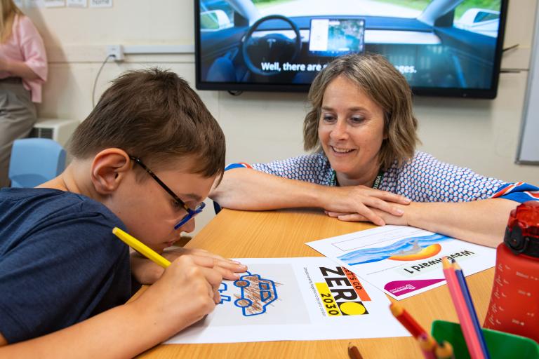 A child colours in a 'Let's Go Zero 2030' colouring page, while a teacher smiles and chats to them.