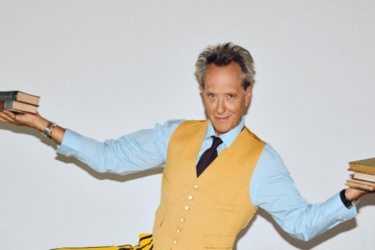Richard E. Grant holds up some books to the camera.