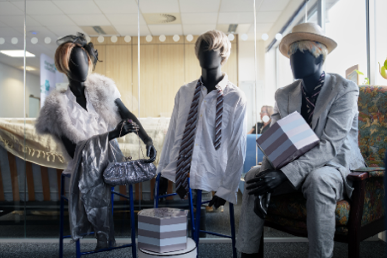 Three mannequins wearing formal clothes sit on chairs on display.