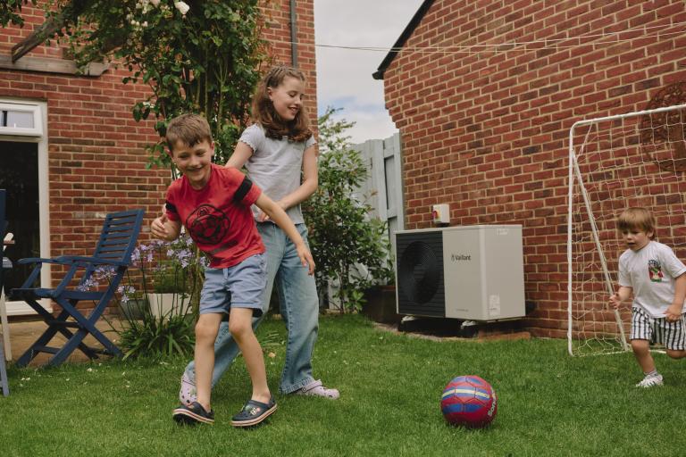 Three children play football in their back garden. A heat pump is visible in the background.