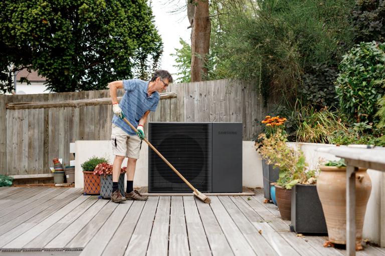 A man sweeps his back garden decking. A heat pump is visible in the background.