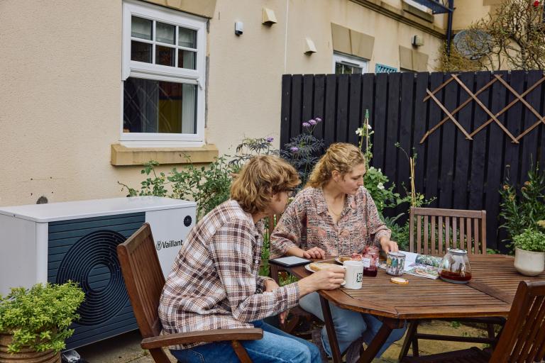 Two women sit at a table in their back garden. A heatpump is seen in the background.
