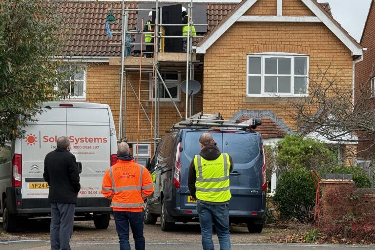 Three men stand outside a house in hi-vis, watching solar panels being installed.